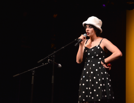 A woman wearing a black dress with white polka-dots and a white hat on a stage singing.